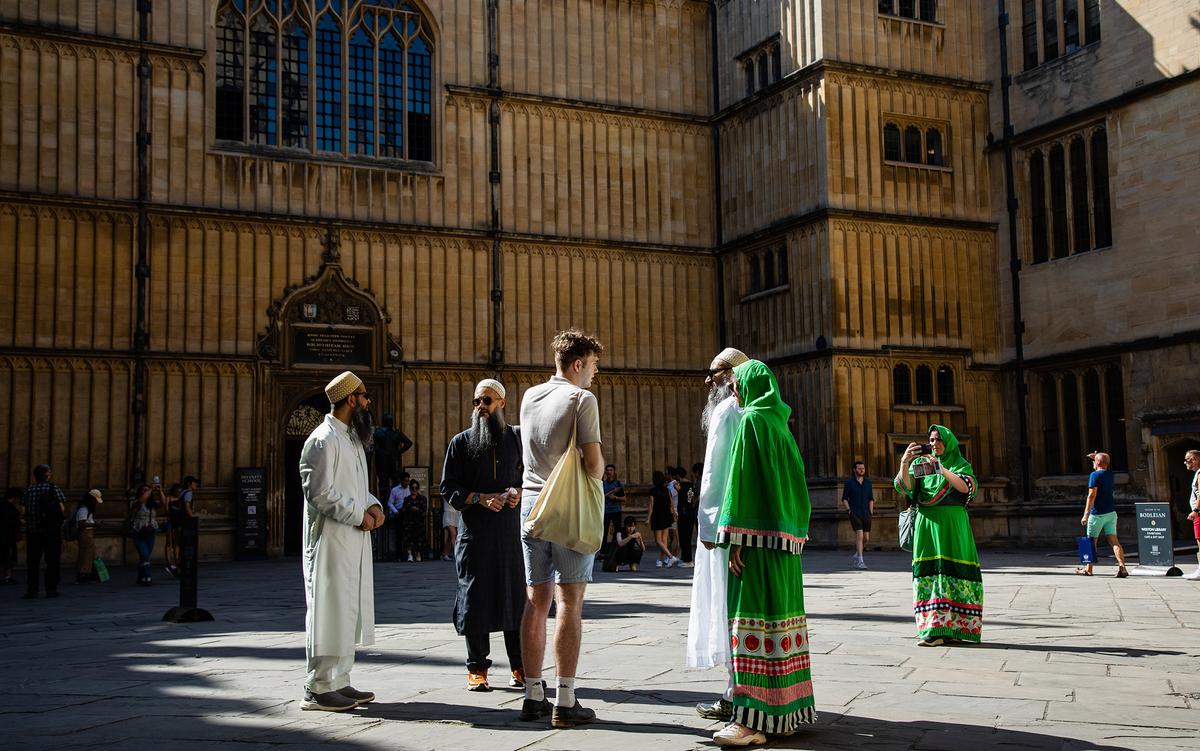 People in traditional and casual clothing conversing outside a historic building with ornate windows. A woman in green takes photos.