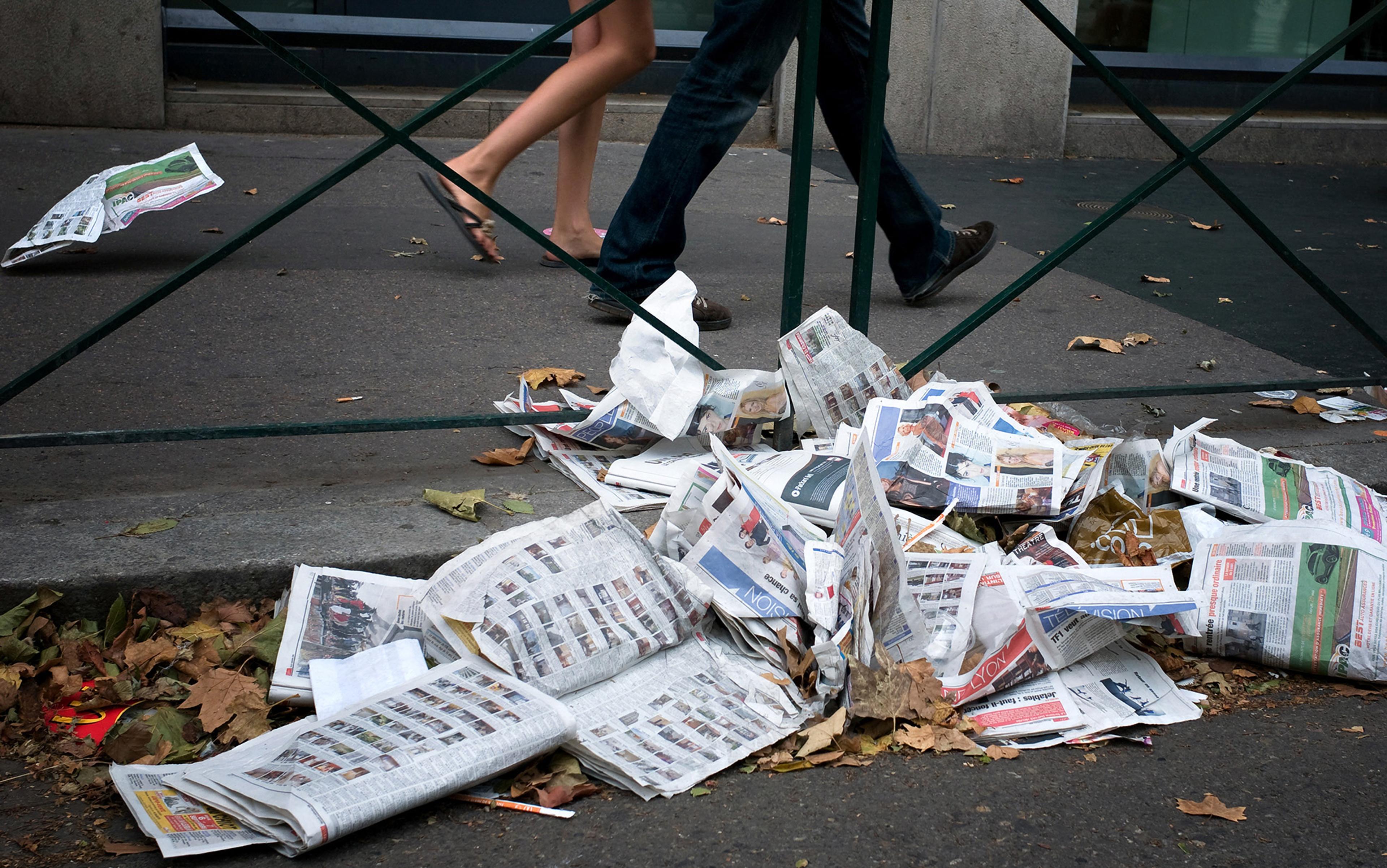 Newspapers and leaves scattered on the pavement, with two people walking past a wrought iron fence in the background.