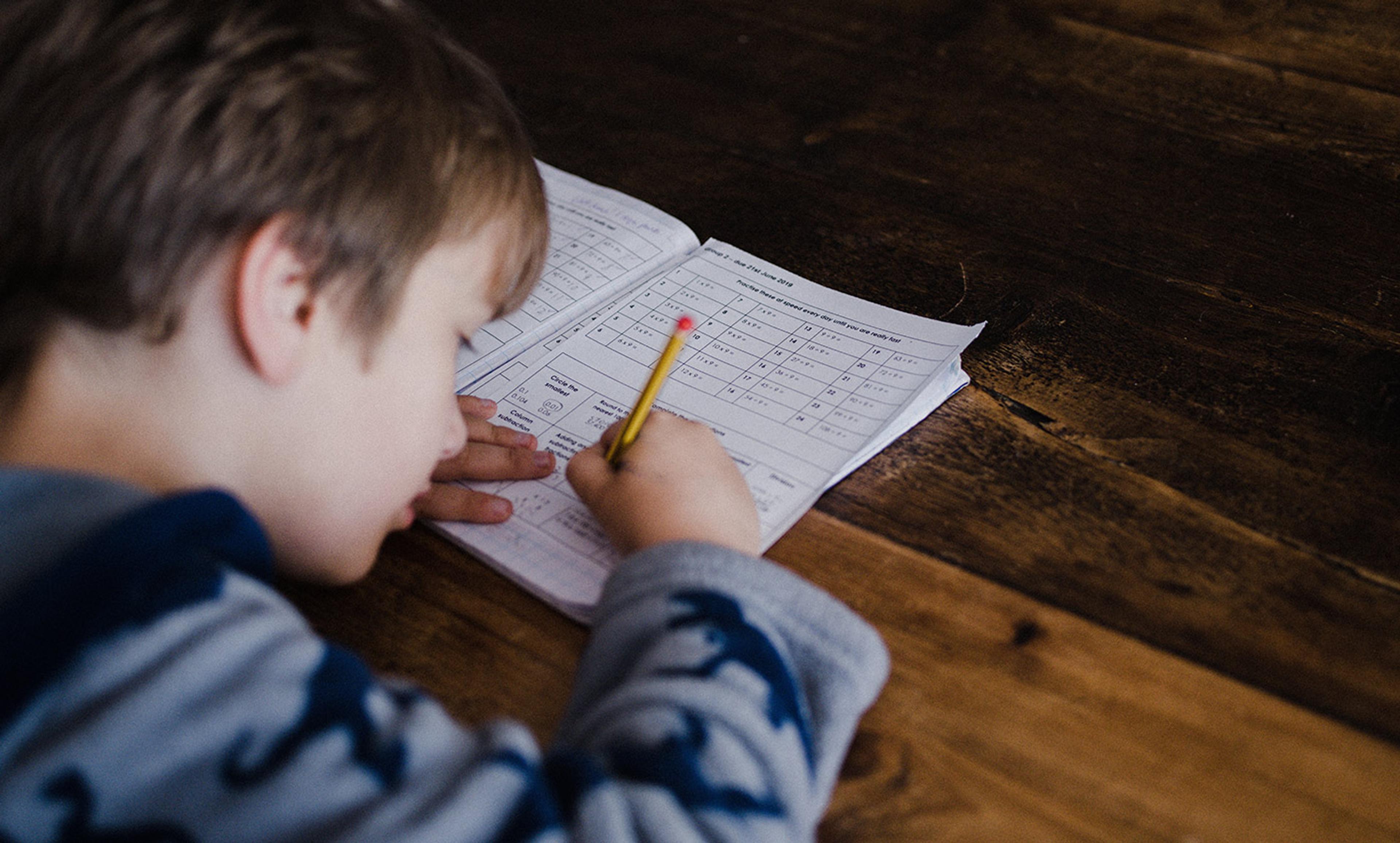 A child writing in a workbook on a wooden table, focused on their task while holding a pencil in their right hand.