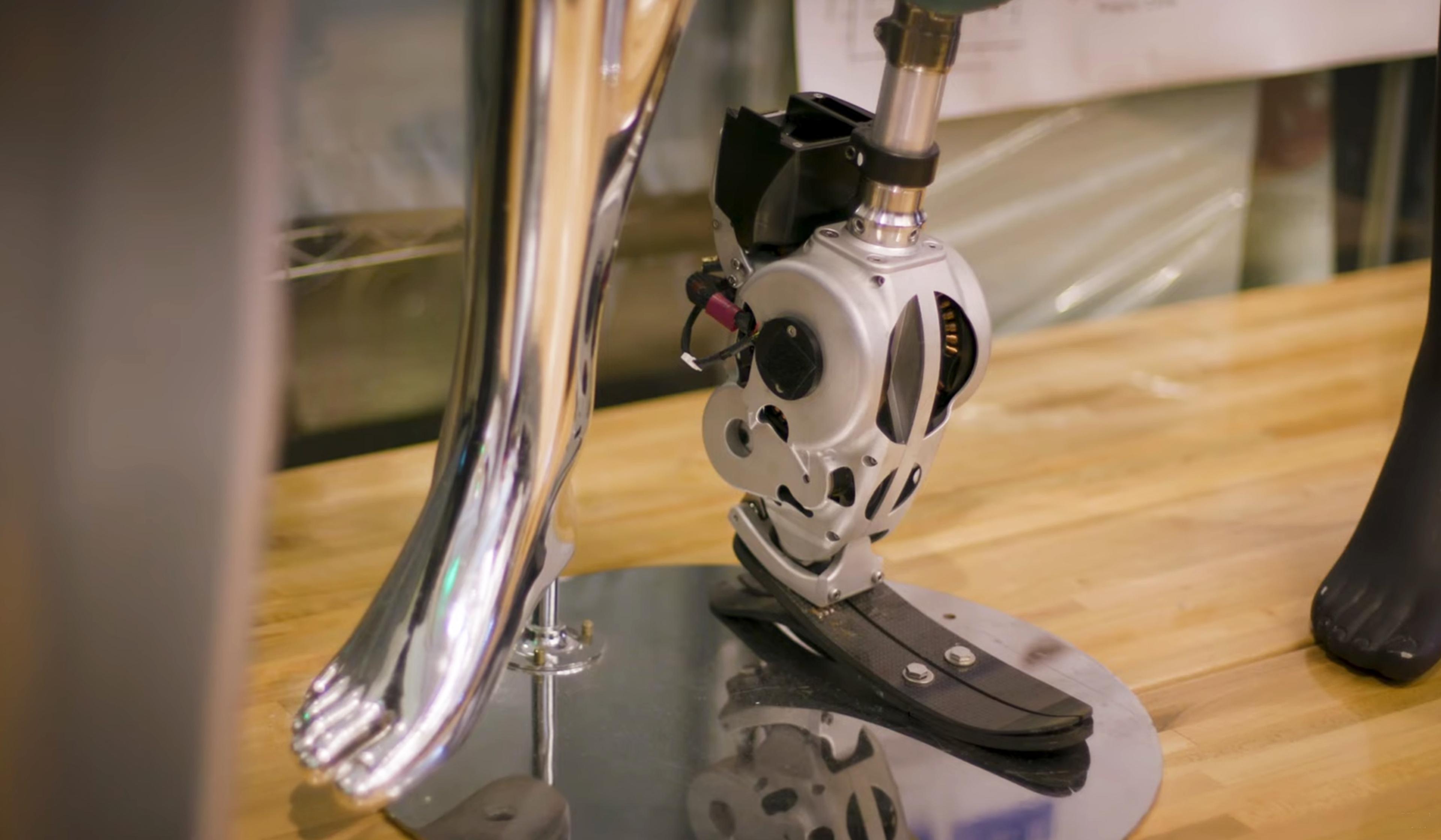 A close-up of a robotic prosthetic leg displayed on a wooden surface, with chrome and black components, reflecting light from surroundings.