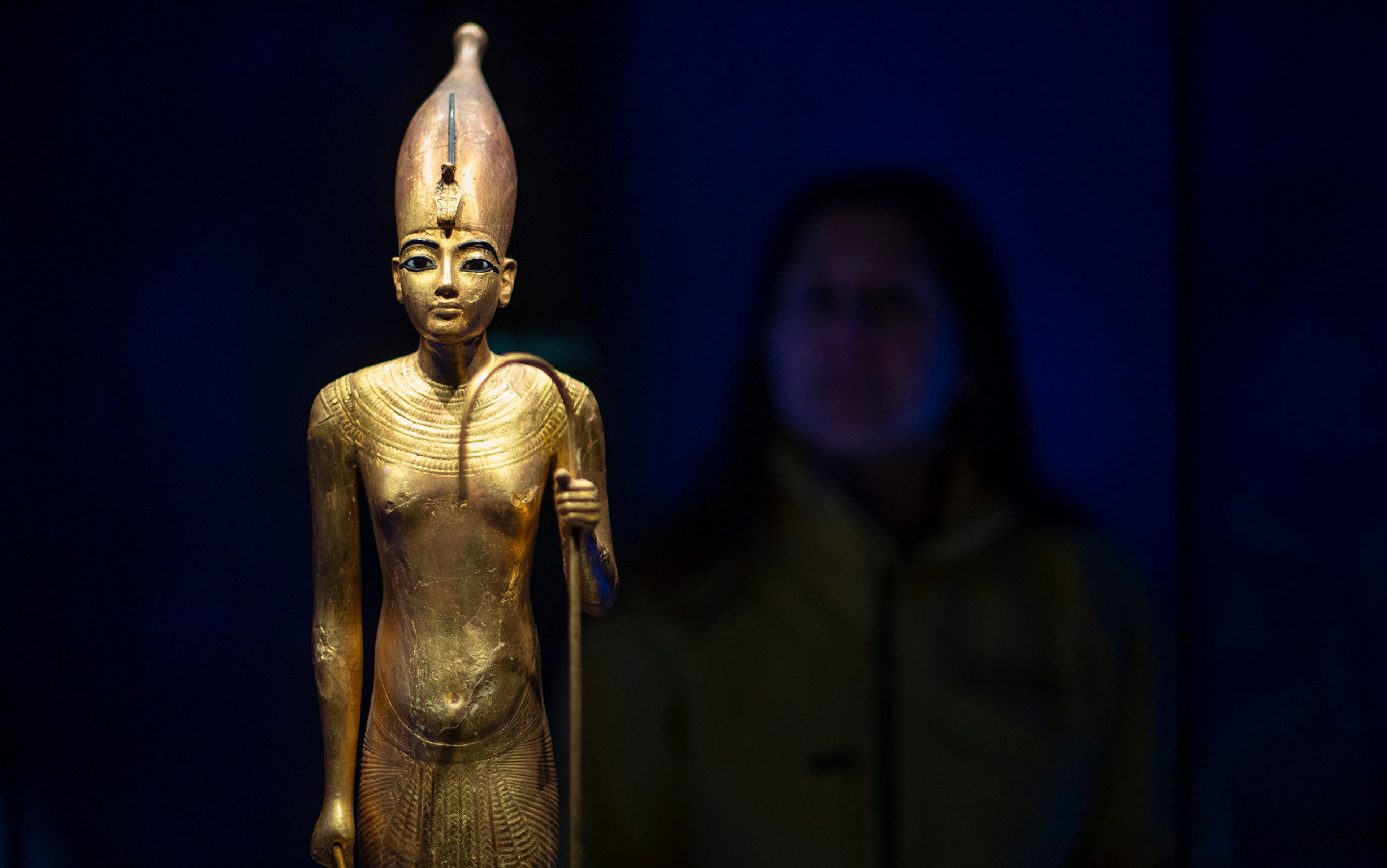 Golden Egyptian statue with intricate details, holding a staff, is displayed in a dark room.