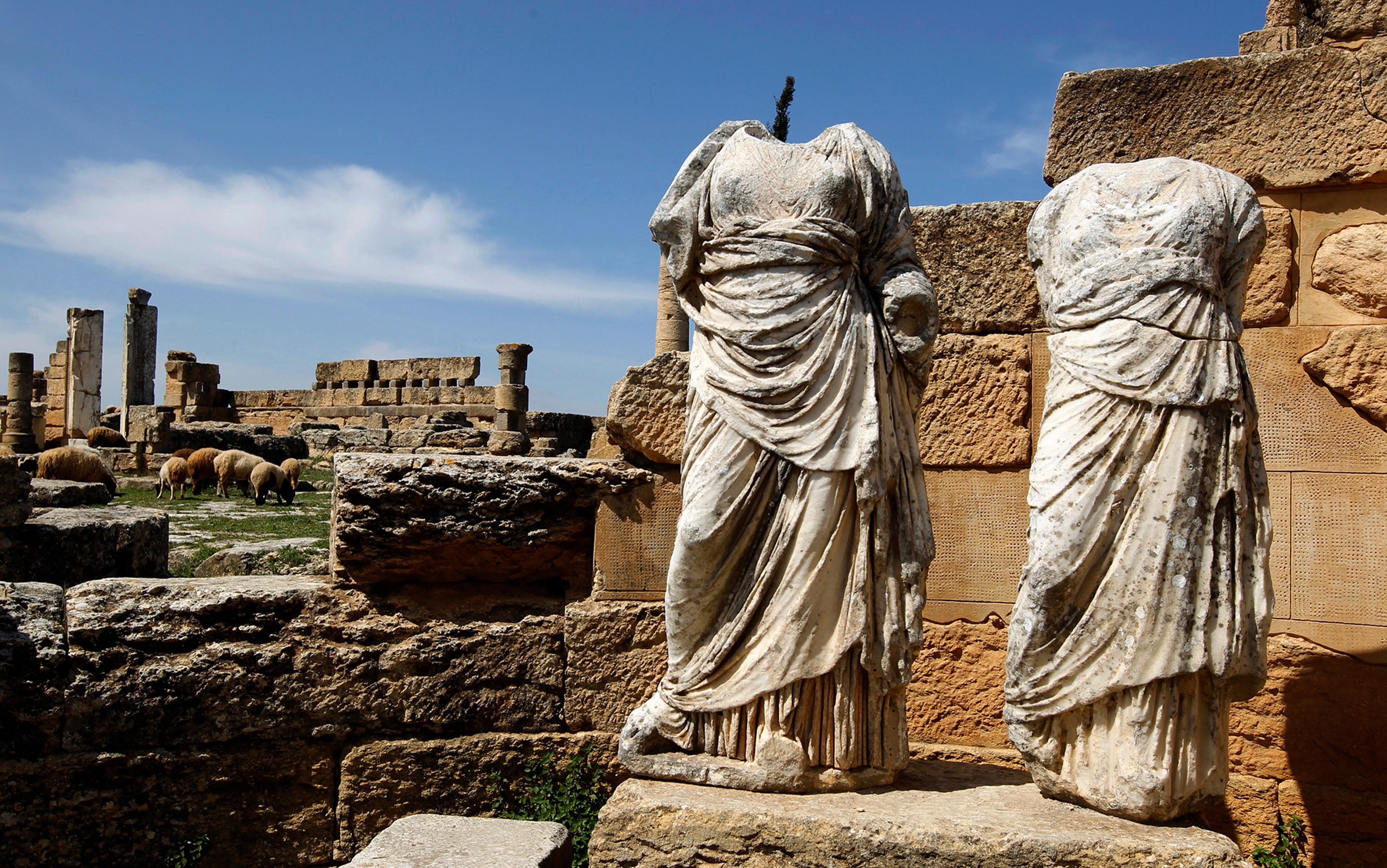 Ancient headless statues amid ruins with grazing sheep in the background under a clear sky.