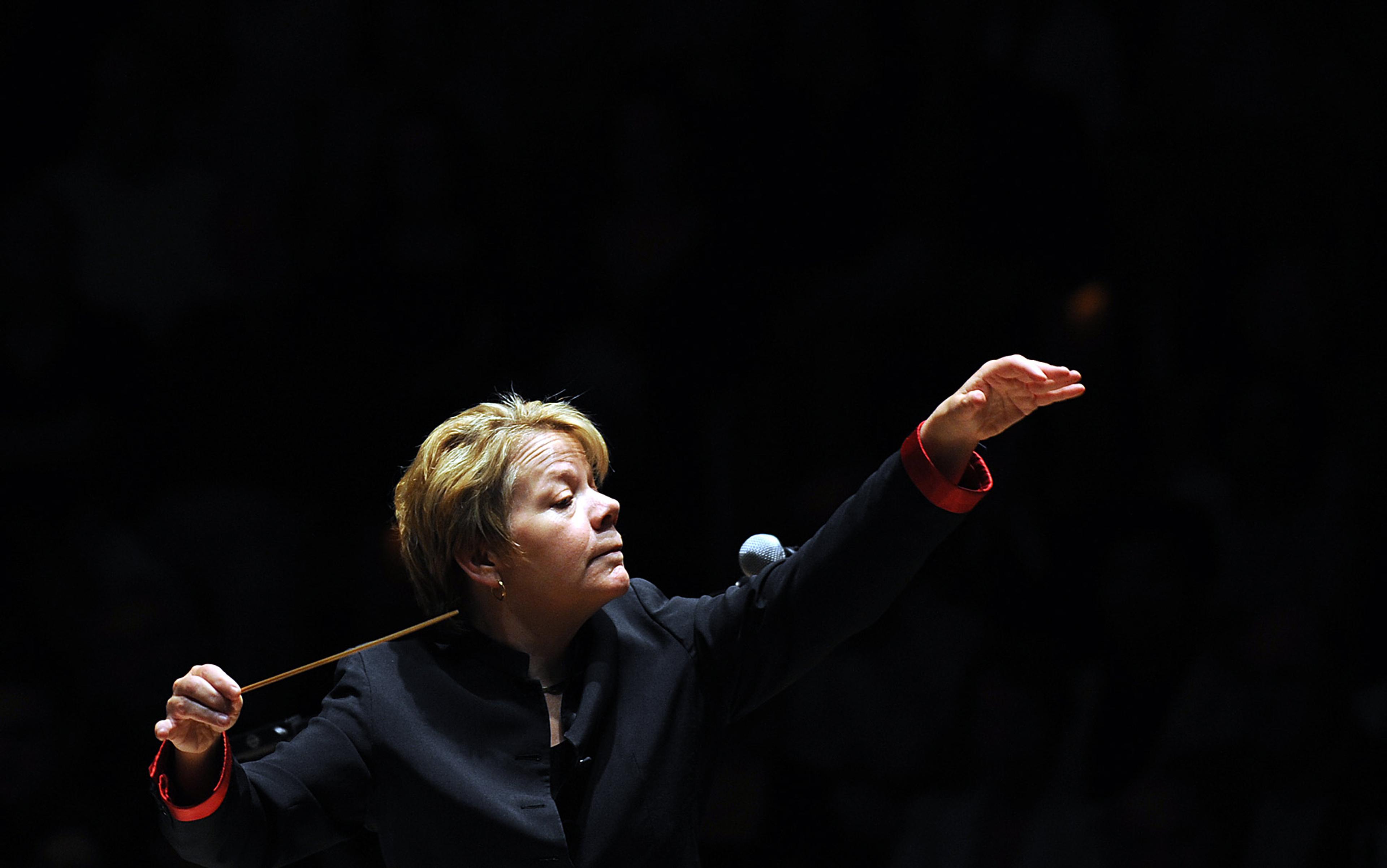 A conductor passionately leads an orchestra, holding a baton in one hand and gesturing with the other, with a focused expression in a dimly lit venue.