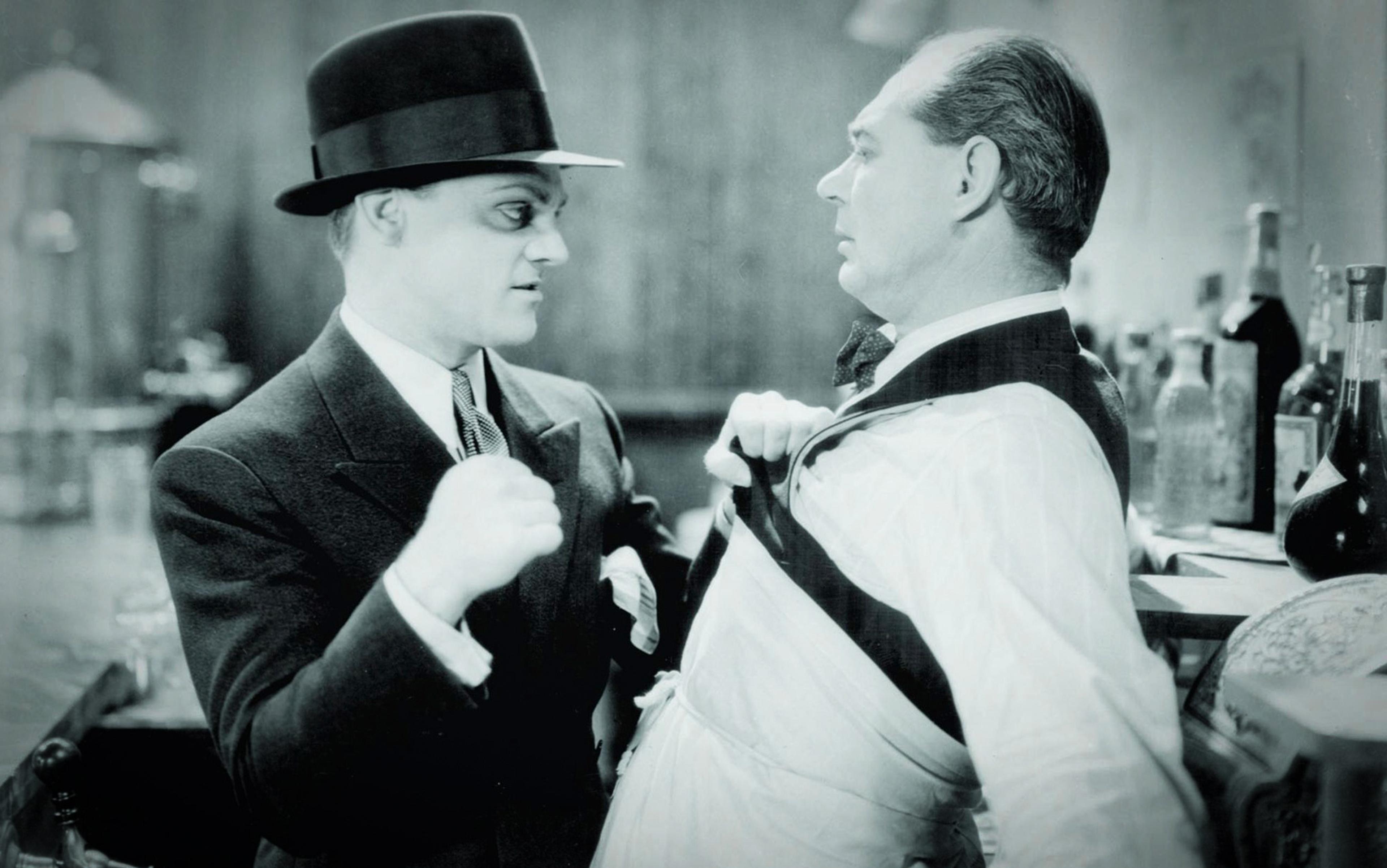 Black-and-white photo of a man in a suit and hat grabbing another man by his collar in front of a bar with bottles.