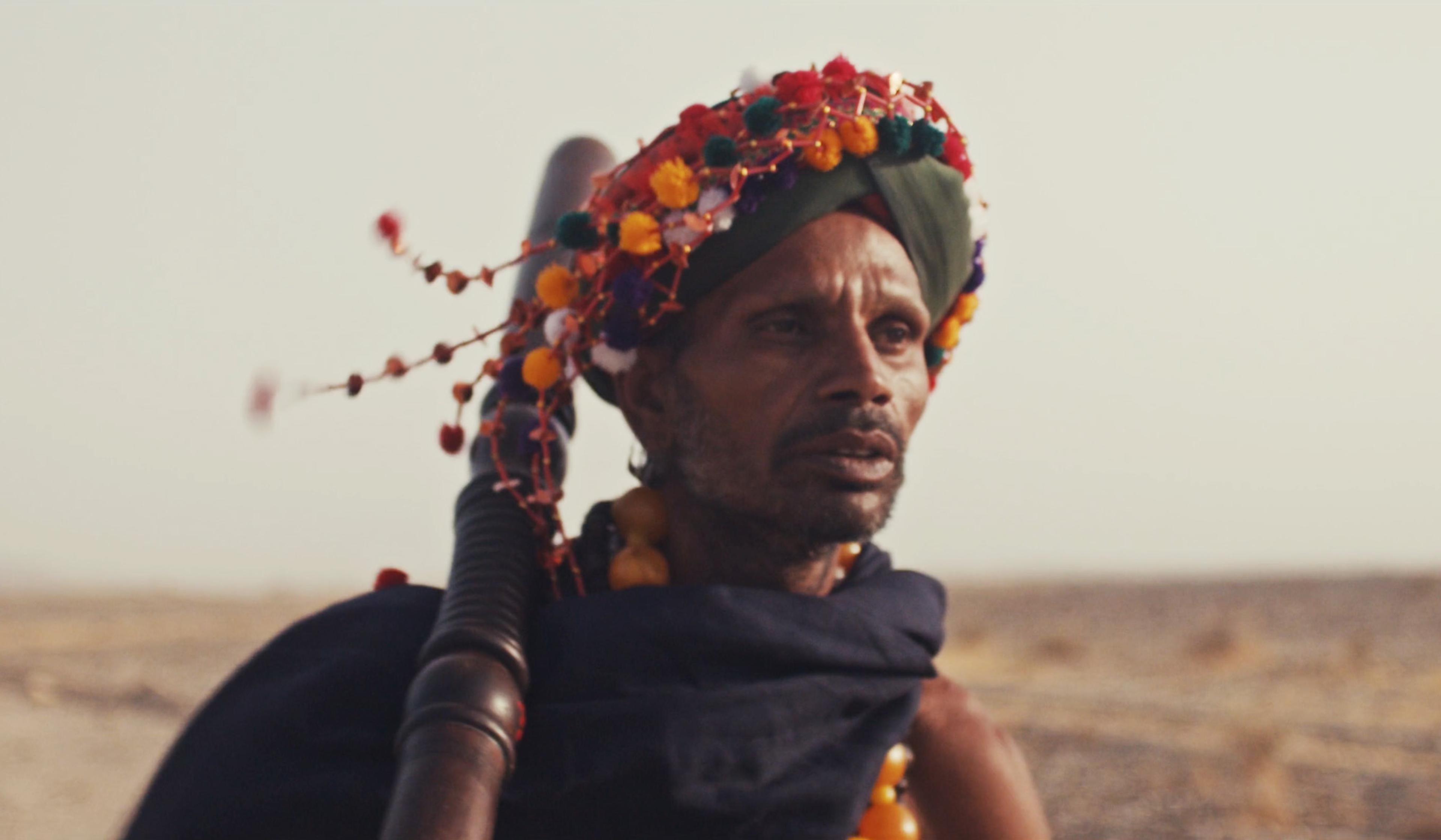 A man in traditional attire with a colourful beaded headdress and a staff over his shoulder, standing outdoors.