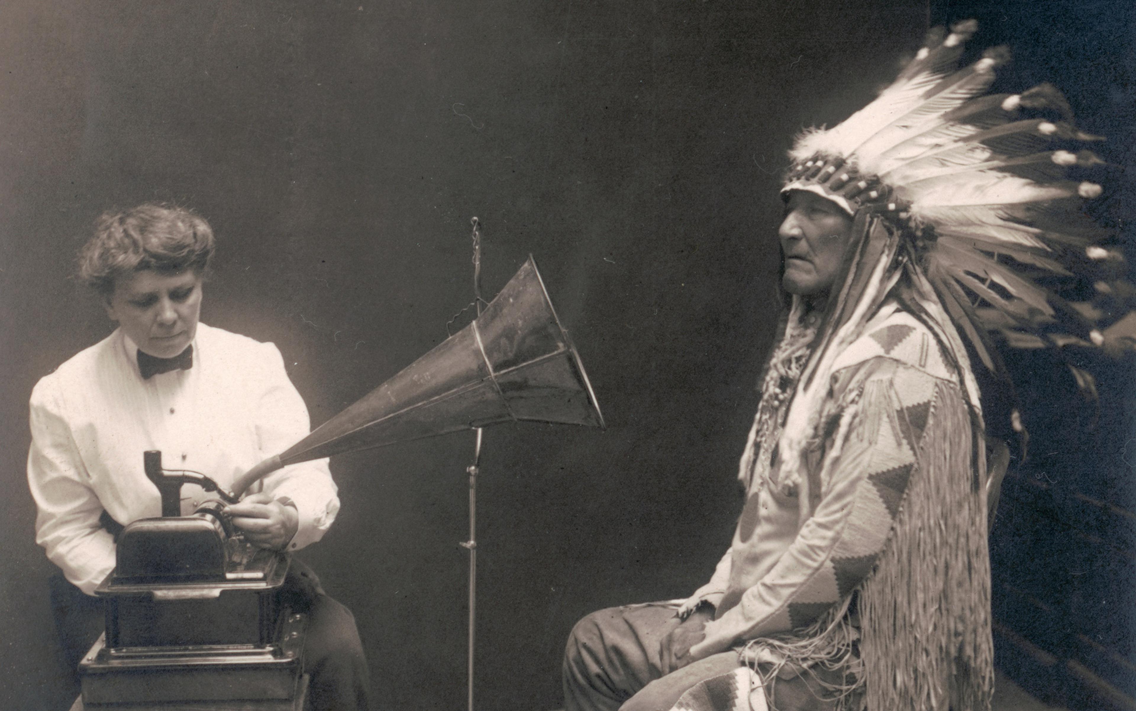 A woman operates an antique phonograph next to a man wearing a traditional Native American headdress in a sepia-toned photograph.