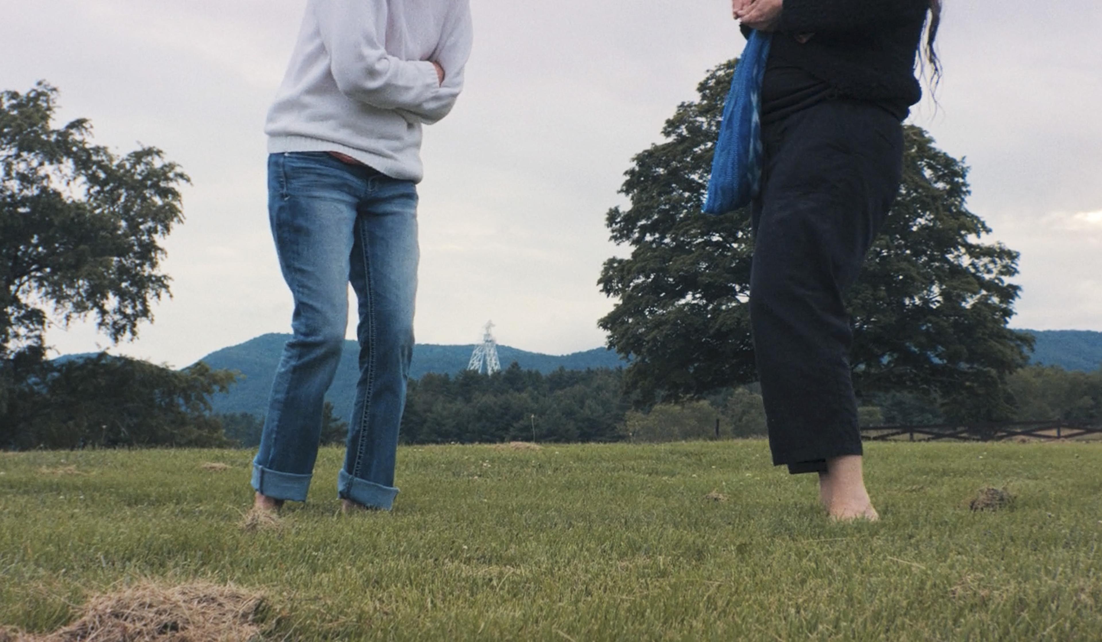 Two people standing barefoot on grass. Only their lower bodies are visible, with hills and trees in the background.