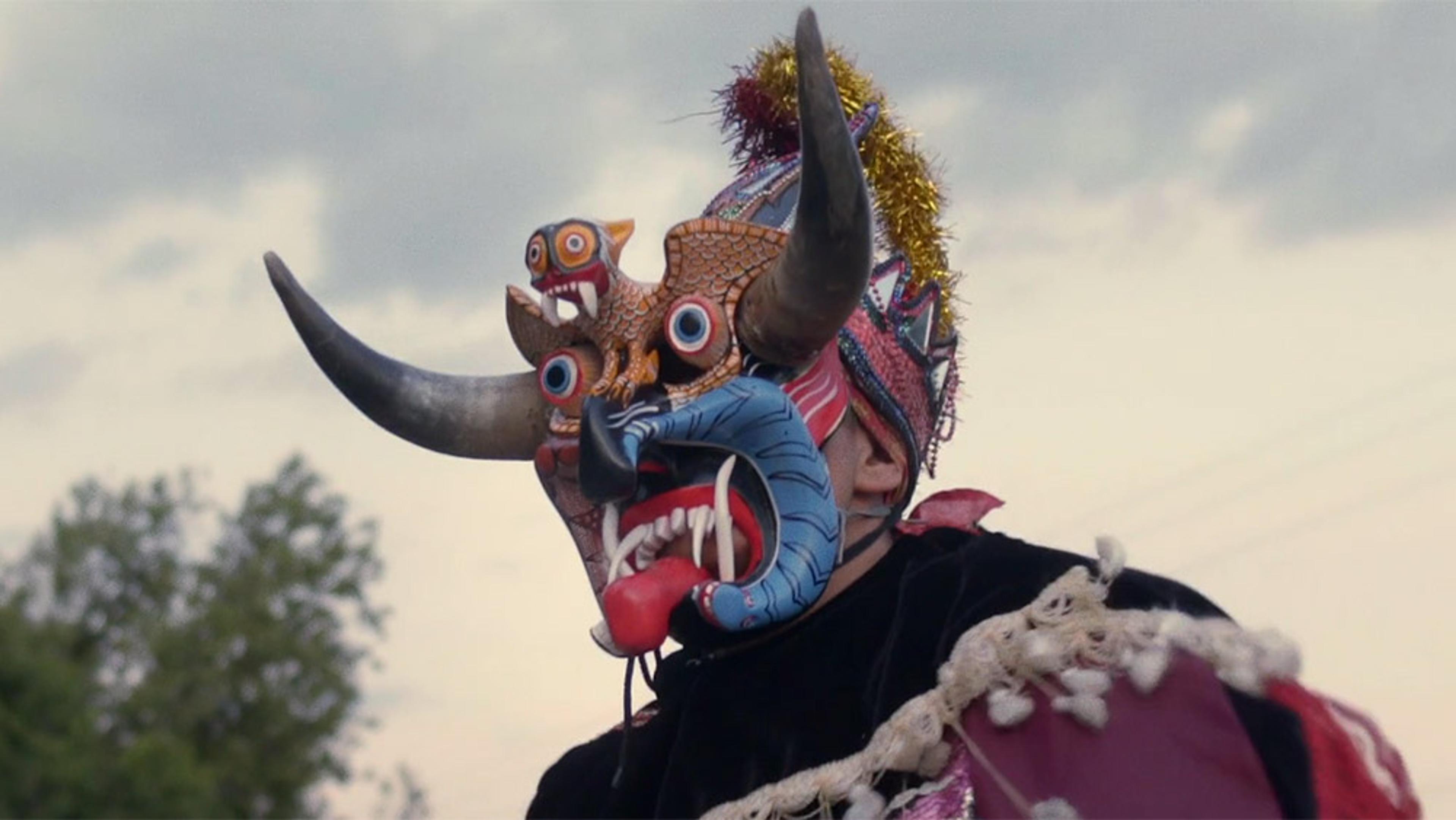 A person wearing a colourful traditional mask with horns, fangs and intricate details against a cloudy sky backdrop.