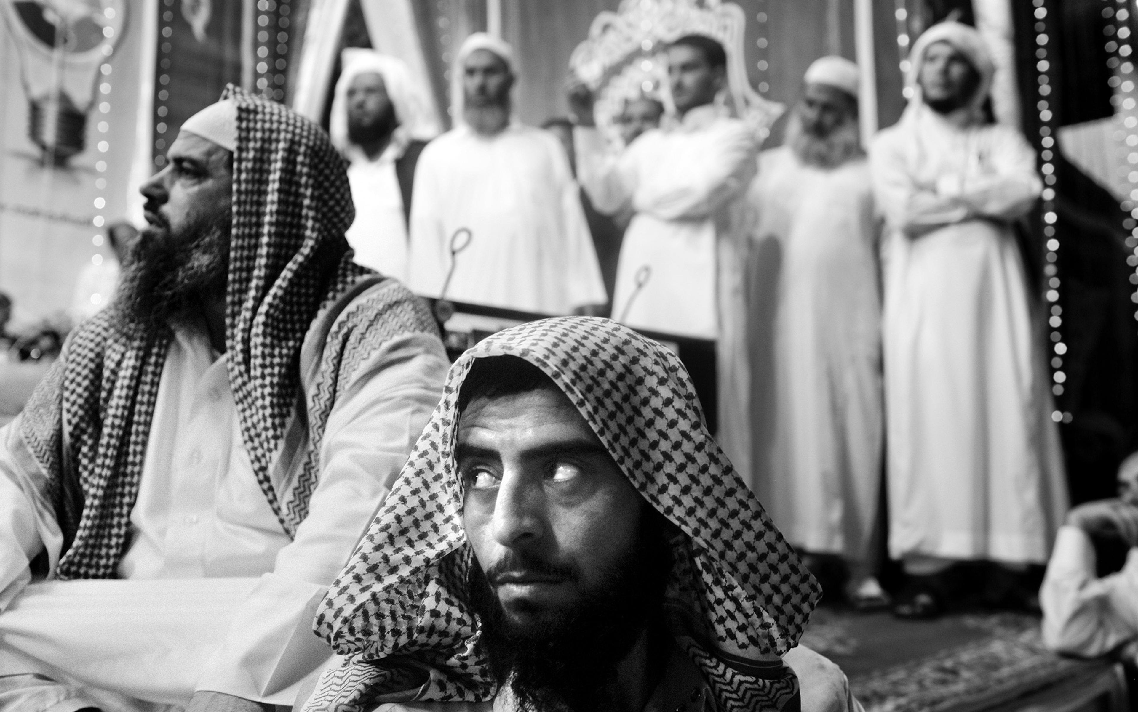 A black and white photo showing a group of men in traditional Middle Eastern attire, with two men seated in the foreground and several standing behind.