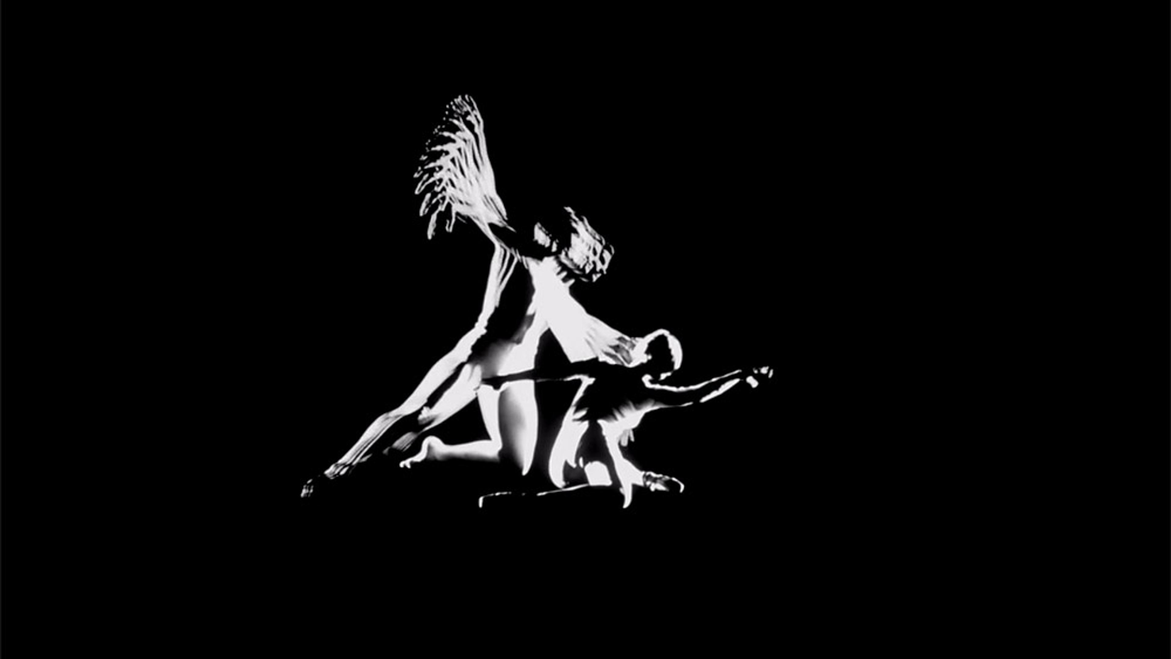 A black and white photo of two dancing figures, one with wings hovering over the other who is kneeling with an arm extended.