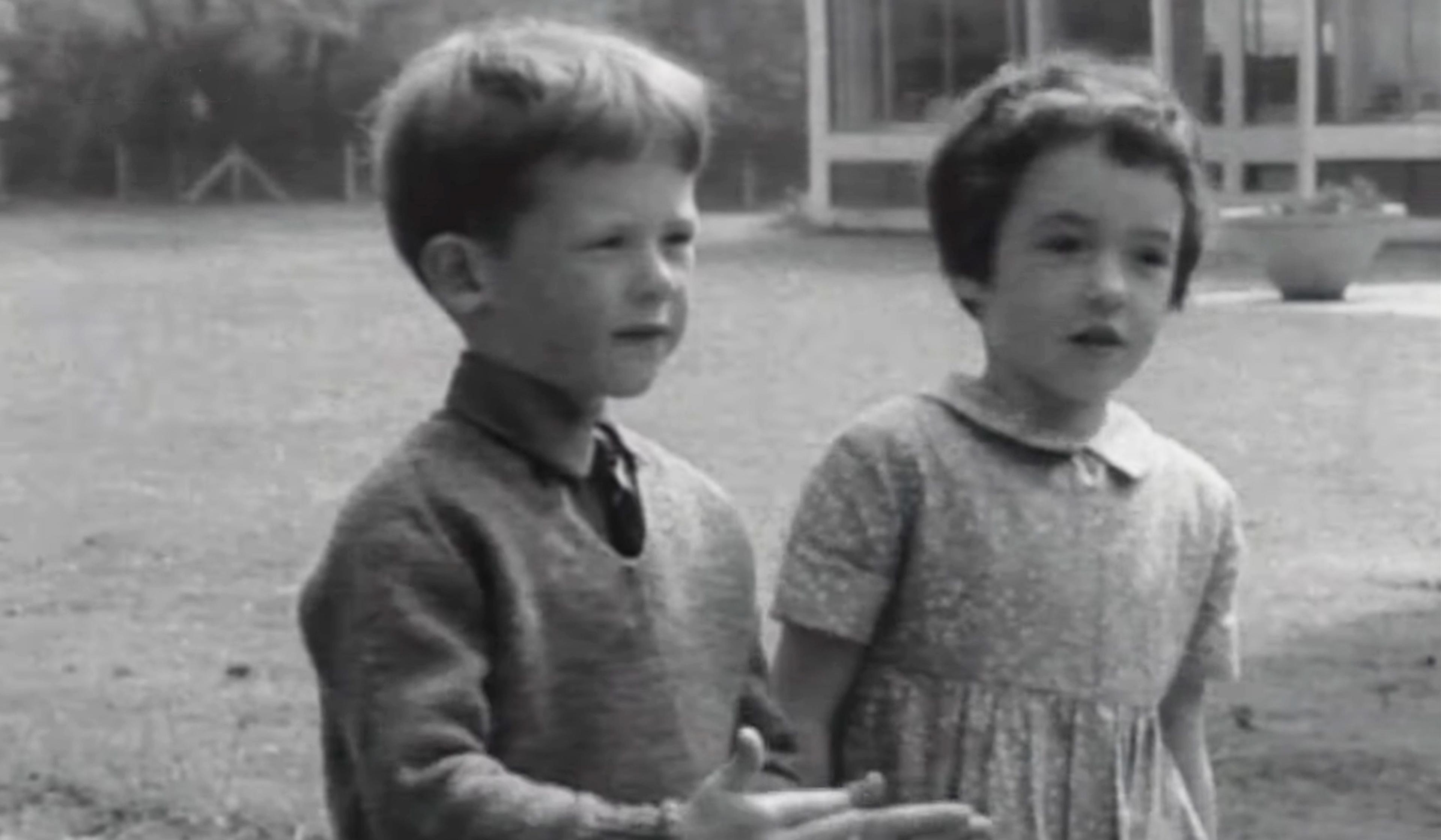 Vintage black-and-white image of two young children standing outside, a boy wearing a jumper and a girl in a dress. They’re in the middle of explaining religion.