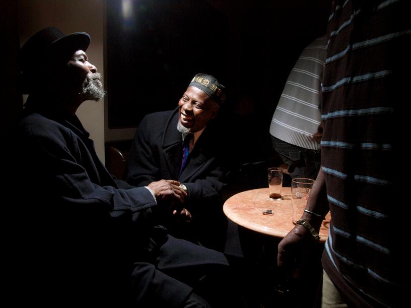 Two men smiling in low light, seated at a round table with drinks. One man is wearing a hat.