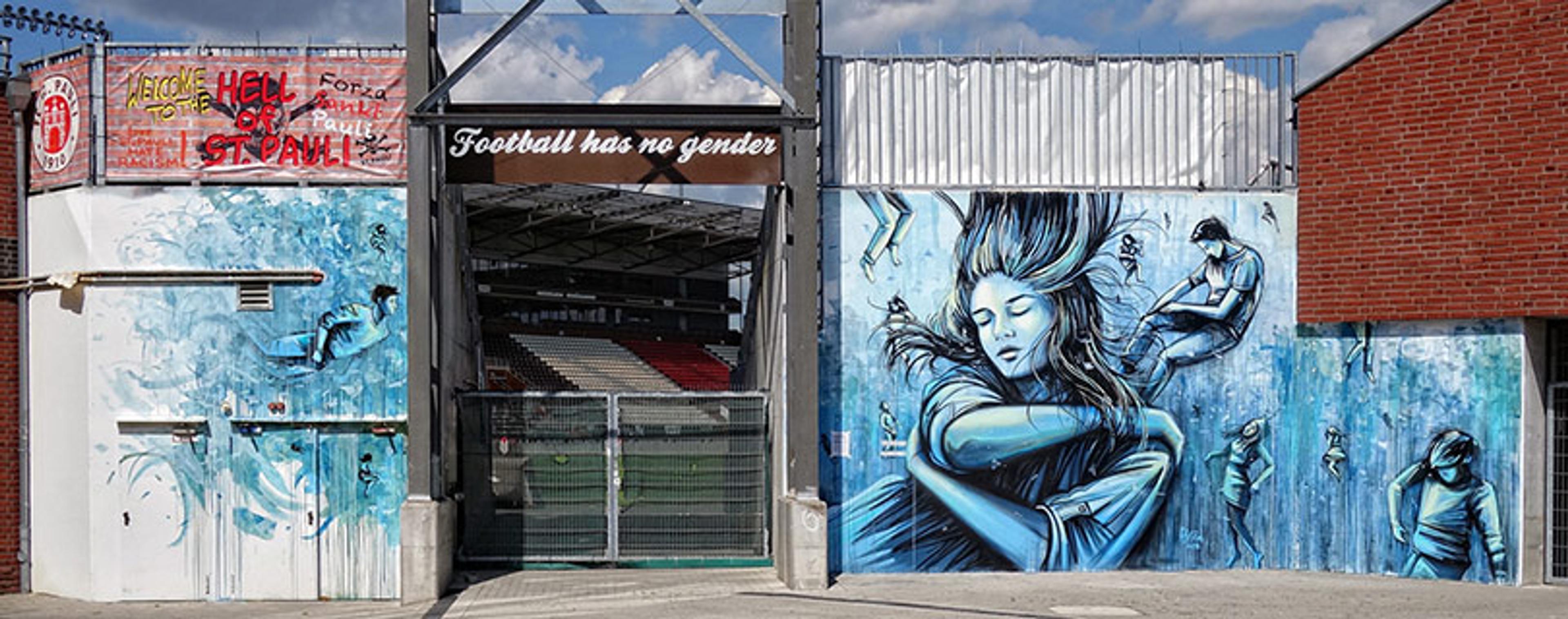 An entrance to a football stadium with a banner reading “Football has no gender”. The surrounding walls feature murals of a woman and other people in shades of blue. Another banner to the left displays “Welcome to the Hell of St. Pauli”.