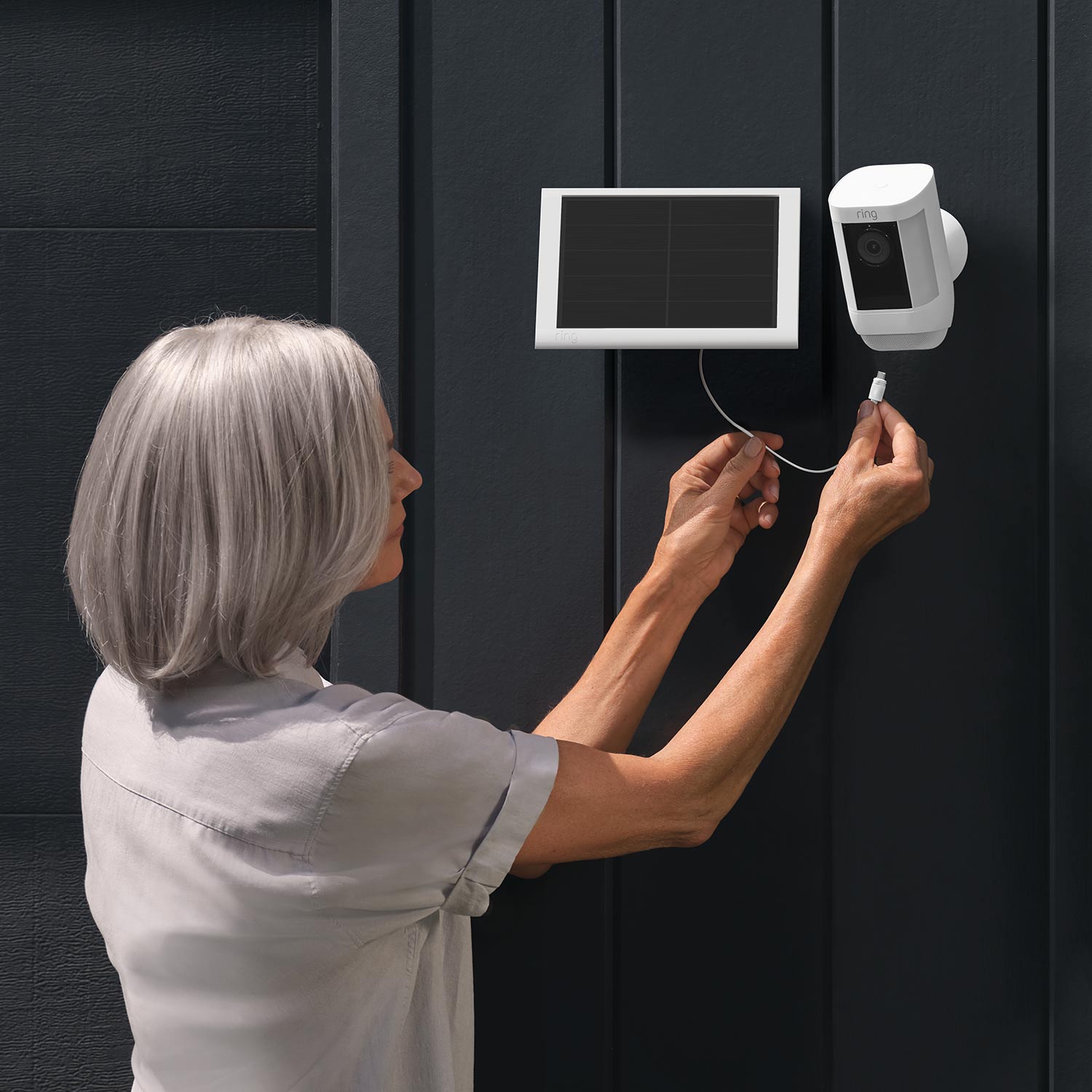 Spotlight Cam Pro - Solar:Spotlight Cam Pro Woman installing cable from solar panel to Spotlight Cam Pro mounted on exterior wall of home.