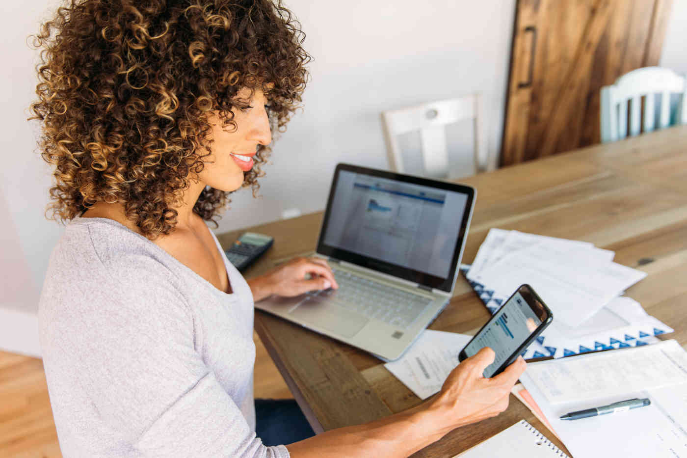 A woman uses a smartphone and computer while reviewing paperwork.