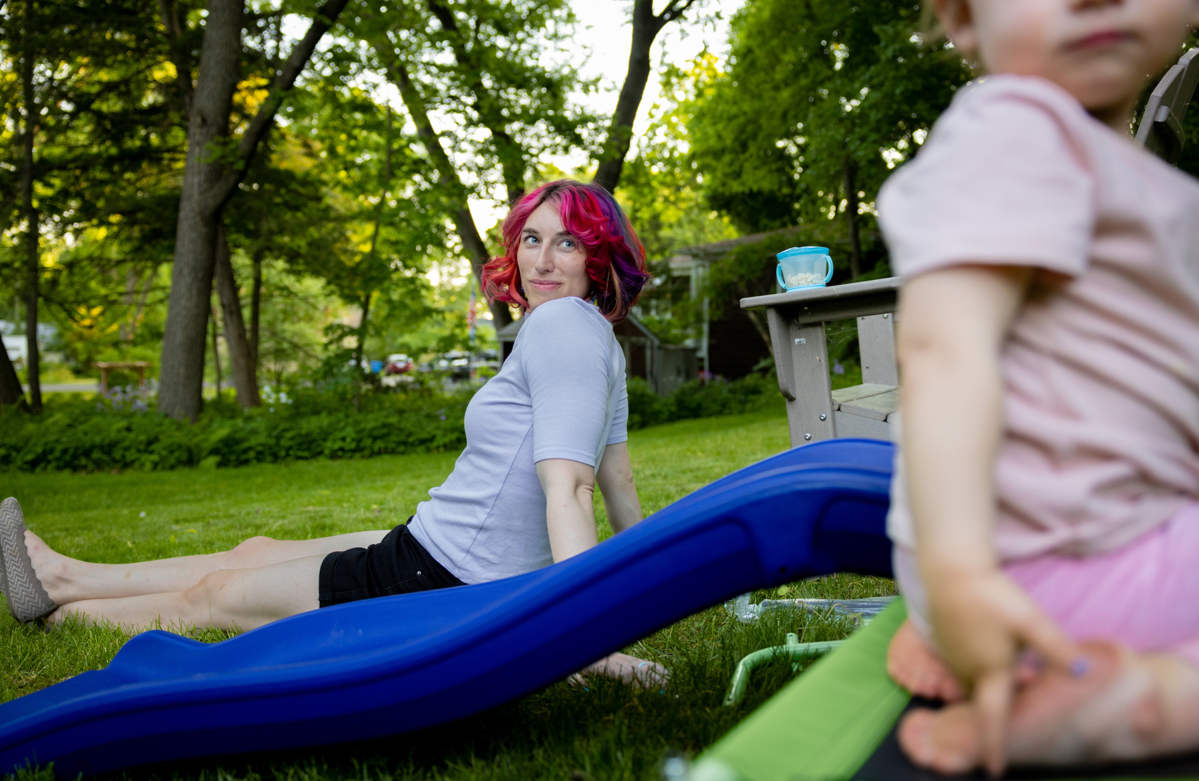 Kyler sits in the grass looking lovingly at her daughter, who’s playing behind her.