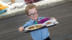 Un petit garçon à lunettes marche fièrement en tenant avec ses deux mains un plateau contenant une grosse assiette de pâtes.