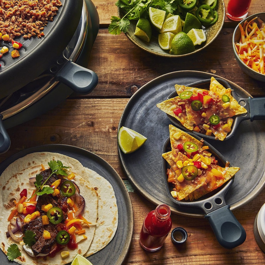 Sur une table en bois, il y a une première assiette avec une tortilla remplie de bavette de bœuf, de maïs, de jalapenos et de coriandre fraîche. Dans une deuxième assiette, il y a des coupelles contenant des nachos à gratiner. Autour, il y a un bol de limes, un bol de fromage, un bol de croustille de maïs et le poêle à raclette sur lequel cuit de la bavette, du maïs et de la PVT.