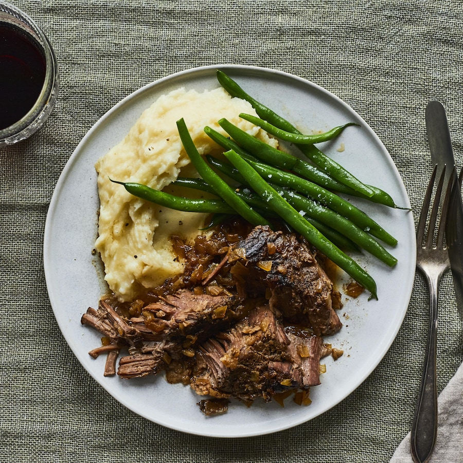 Du rôti de palette, des pommes de terre pilées et des haricots verts dans une assiette sur une nappe, avec un couteau et une fourchette. 