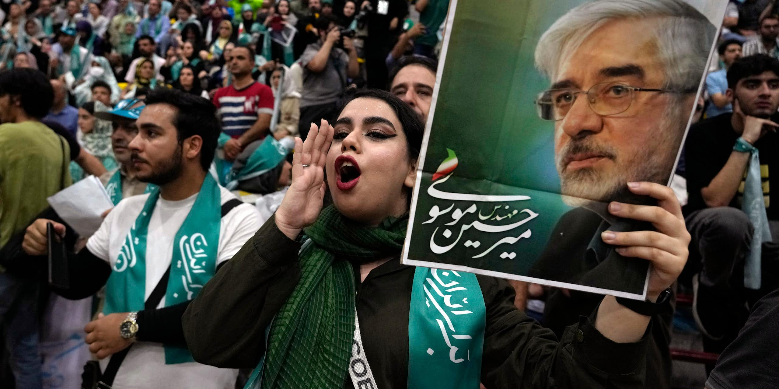 A large crowd of Iranian supporters of Masoud Pezeshkian gather in Tehran; in the foreground, a young woman shouts her endorsement while holding up a campaign sign.