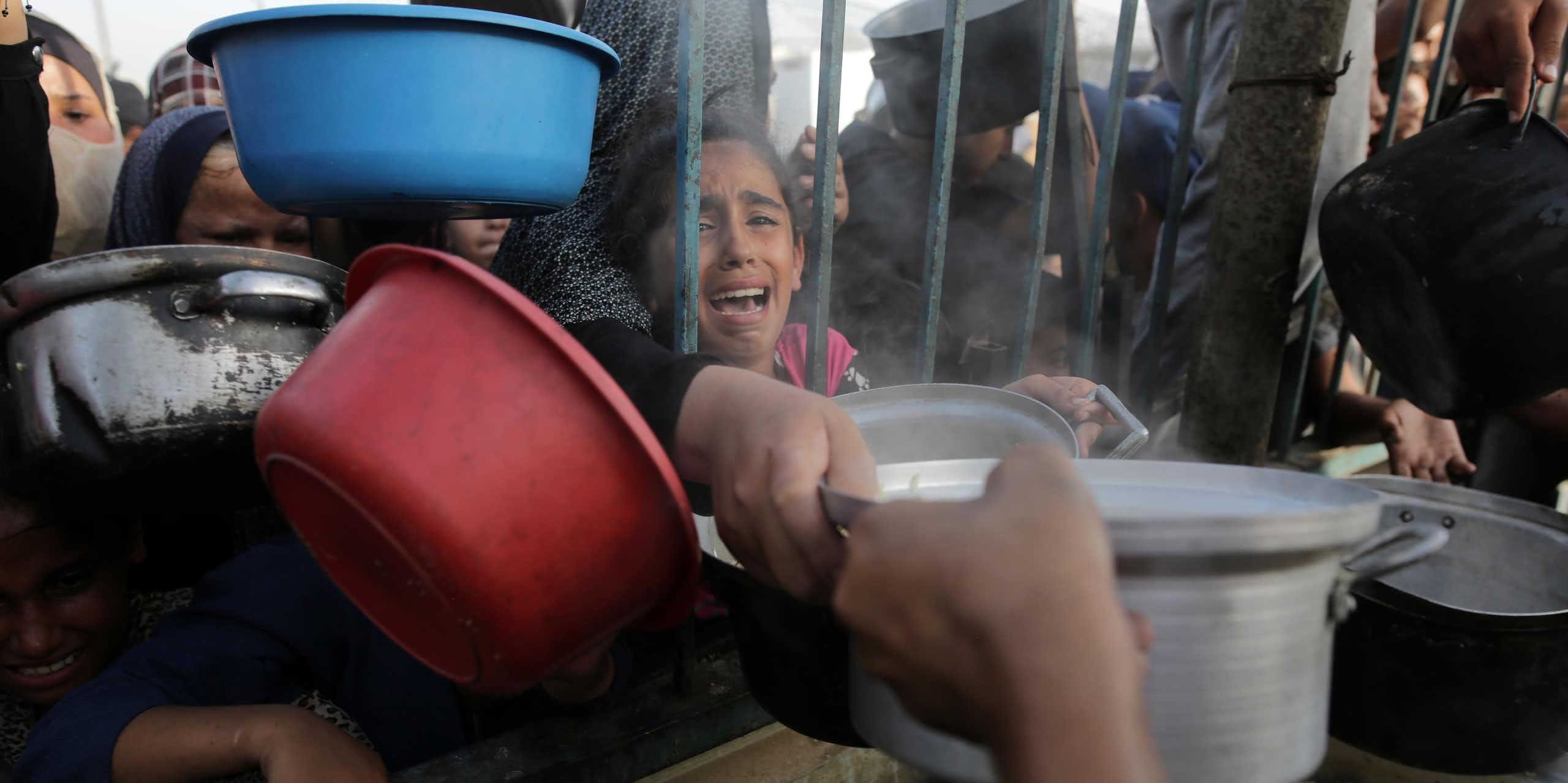 A young girl cries as she reaches through bars for food, as other people clamor around her with bowls.