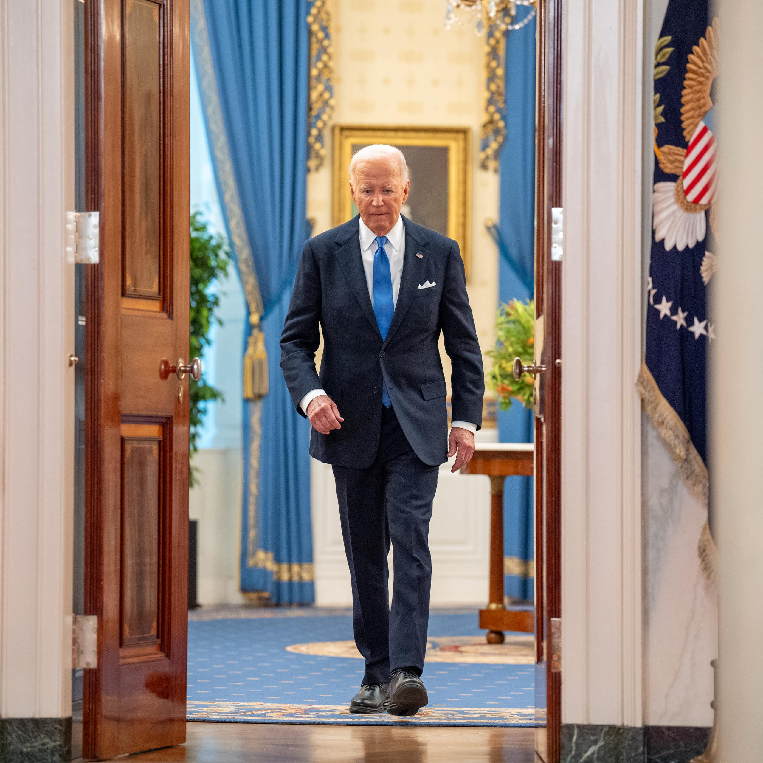 A white man with white hair, a navy suit and a blue tie walks through open doors, with an American flag on one side of it.