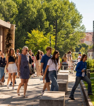 Students walking and socializing outside a university campus building on a sunny day.