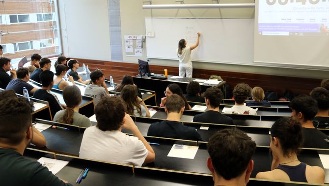 Pla general d'una aula d'universitat amb estudiants d'esquenes mirant les indicacions d'una membre del tribunal