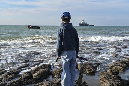 Zuwanderung: A boy stands watching as migrants sit on board a smugglers' boat in order to attempt crossing the English channel off the beach of Audresselles, northern France on October 25, 2024. Two people died while attempting to cross the Channel from northern France to Britain early October 23, 2024, French authorities said, adding that around 50 people more had been rescued from a sinking boat. (Photo by Bernard BARRON / AFP) (Photo by BERNARD BARRON/AFP via Getty Images)