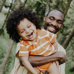 A Black man smiling holding a Black school-age child in his arms who is laughing.