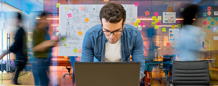 A professional uses his laptop in front of a board filled with ideas on post-it notes while his colleagues busily move around behind him.