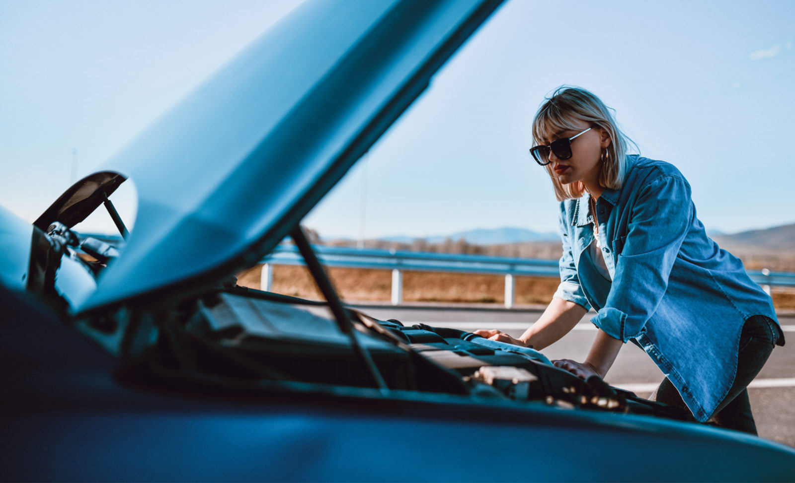 A blond woman wearing sunglasses and a blue collared shirt stands over the exposed hood of her car.