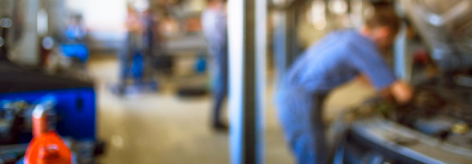 A blurry image of a male mechanic inspecting the exposed under hood of a car in an auto shop.