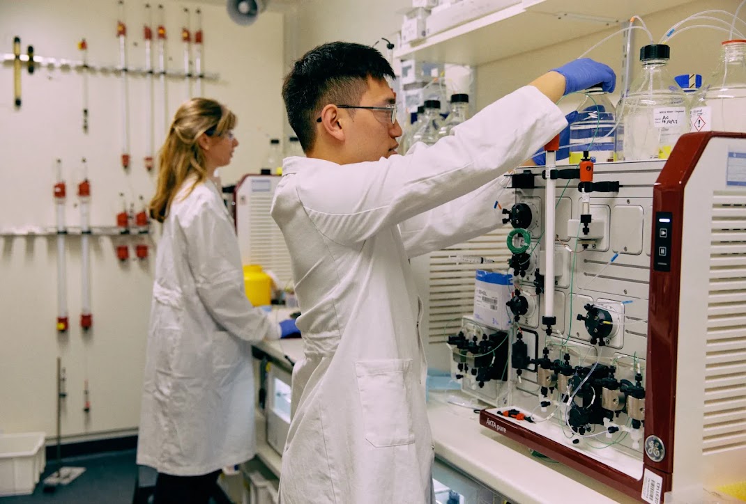 Two scientists performing a complex-looking task in a laboratory. 
They are surrounded by equipment.