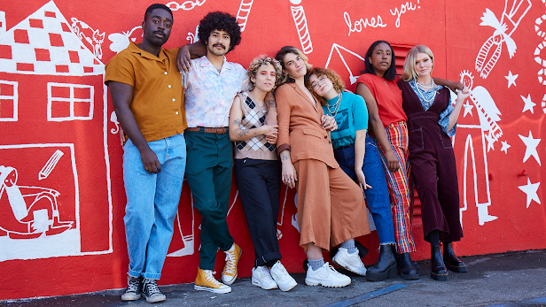 A group of seven people wearing colorful outfits stand outside in front of a red wall with white illustrations