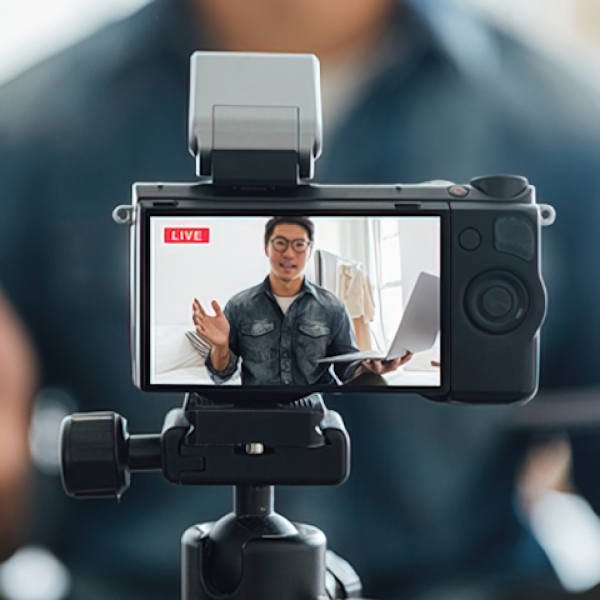 An Asian man records himself and is seen wearing glasses and holding a laptop in the camera's viewfinder