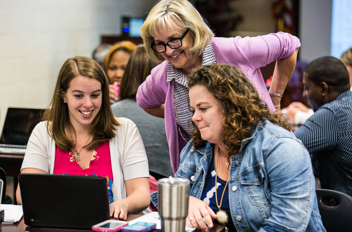 Three educators smile while looking at a laptop. They are in a conference room filled with people sitting at tables working on laptops