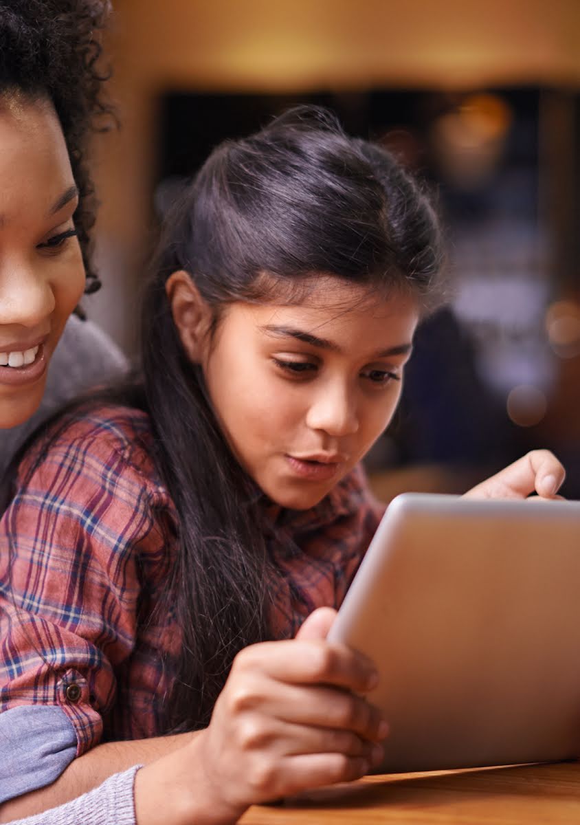 Uma mulher sorridente e uma menina curiosa olham juntas para a tela de um tablet.