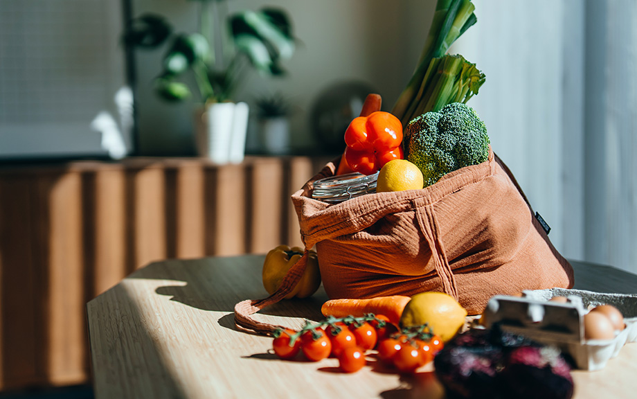 Photo of groceries including peppers, broccoli, lemons and eggs, on a kitchen table in a reusable shopping bag.