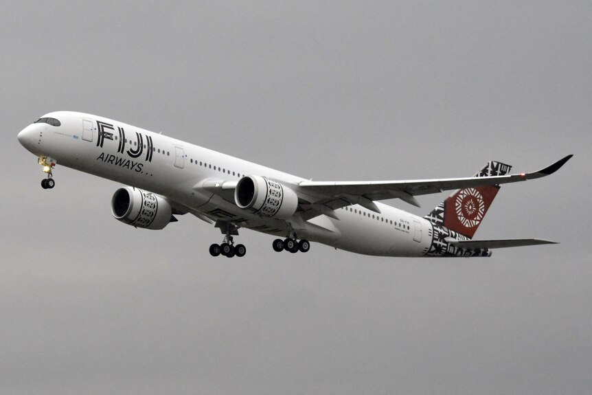 A Fiji Airways plane in flight, with a grey cloudy sky in the background.