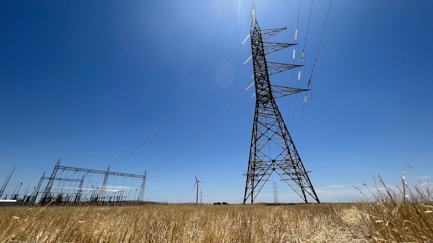 Picture from the ground looking up at a high-voltage transmission tower, with wind turbines in the background