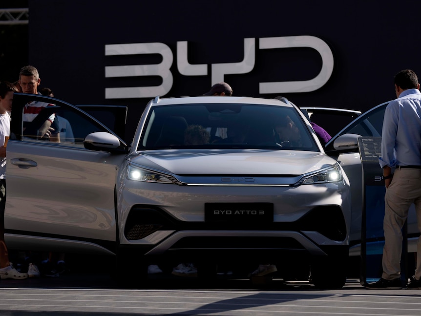 People stand around a car with all its doors open with a sign in beg letters on the wall behind reading BYD