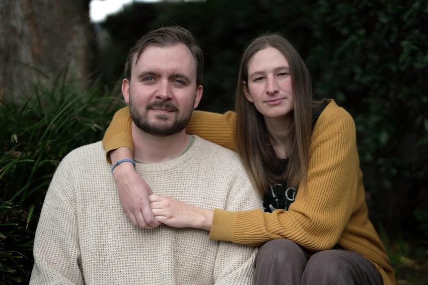 A man and woman sit outdoors looking at the camera with a neutral expression. The woman has her arm around him.