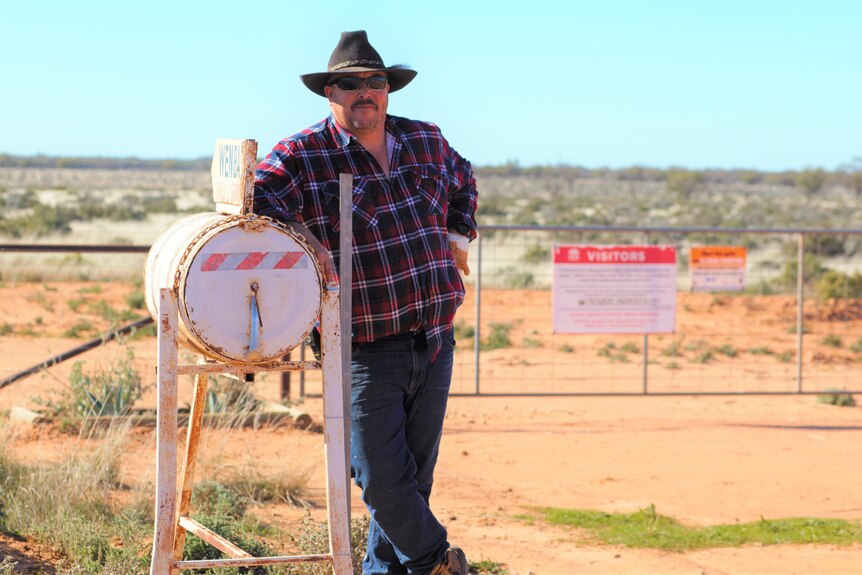 A man wearing a hat stands at a gated entrance to a rural property.