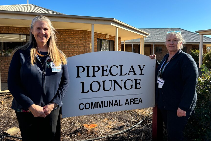 two women stand either side of a sign reading pipeclay lounge communal area on a sunny day with blue skies