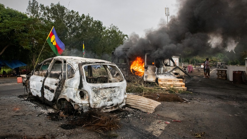 A burnt-out car sits next to a burning car with flames and black smoke pouring from it on a motorway surrounded by green trees