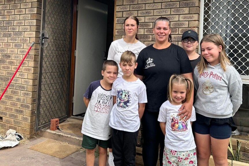 A mum stands with her six young children outside a brown brick building near a wire-mesh door and window, in daylight.