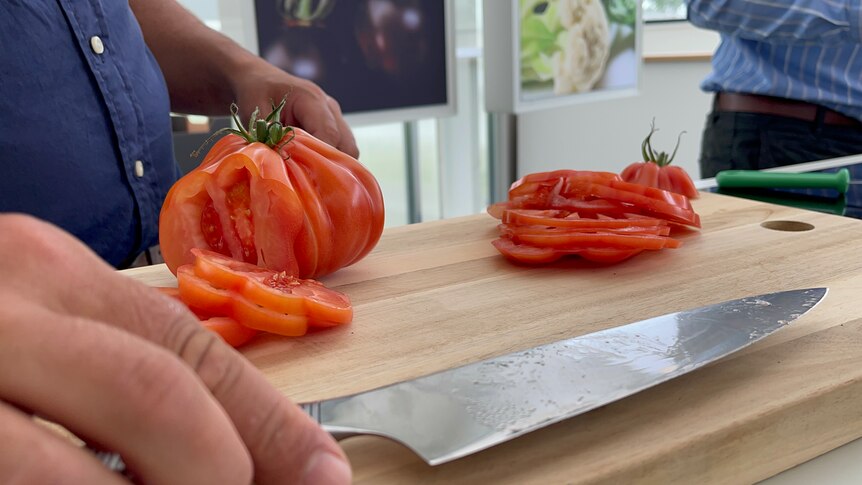 A large oddly shaped tomato has had a few slices cut from it, it's on a wooden board next to a knife 