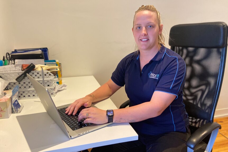 A woman with blonde hair wearing a navy blue shirt sits at her desk typing on her computer