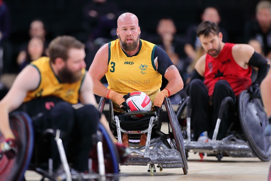 An Australian wheelchair rugby player holds the ball in his lap while he sits in his chair on the court during a game.